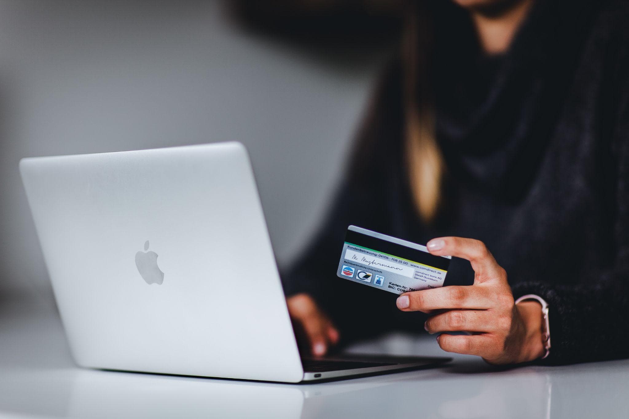 A hand holding a debit card as someone looks at a laptop